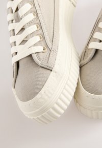 Beige canvas sneakers met een gestructureerde bovenkant, witte veters en een geribbelde rubberen zool. Accentueert met lichtgrijze stiksels en oogjes.