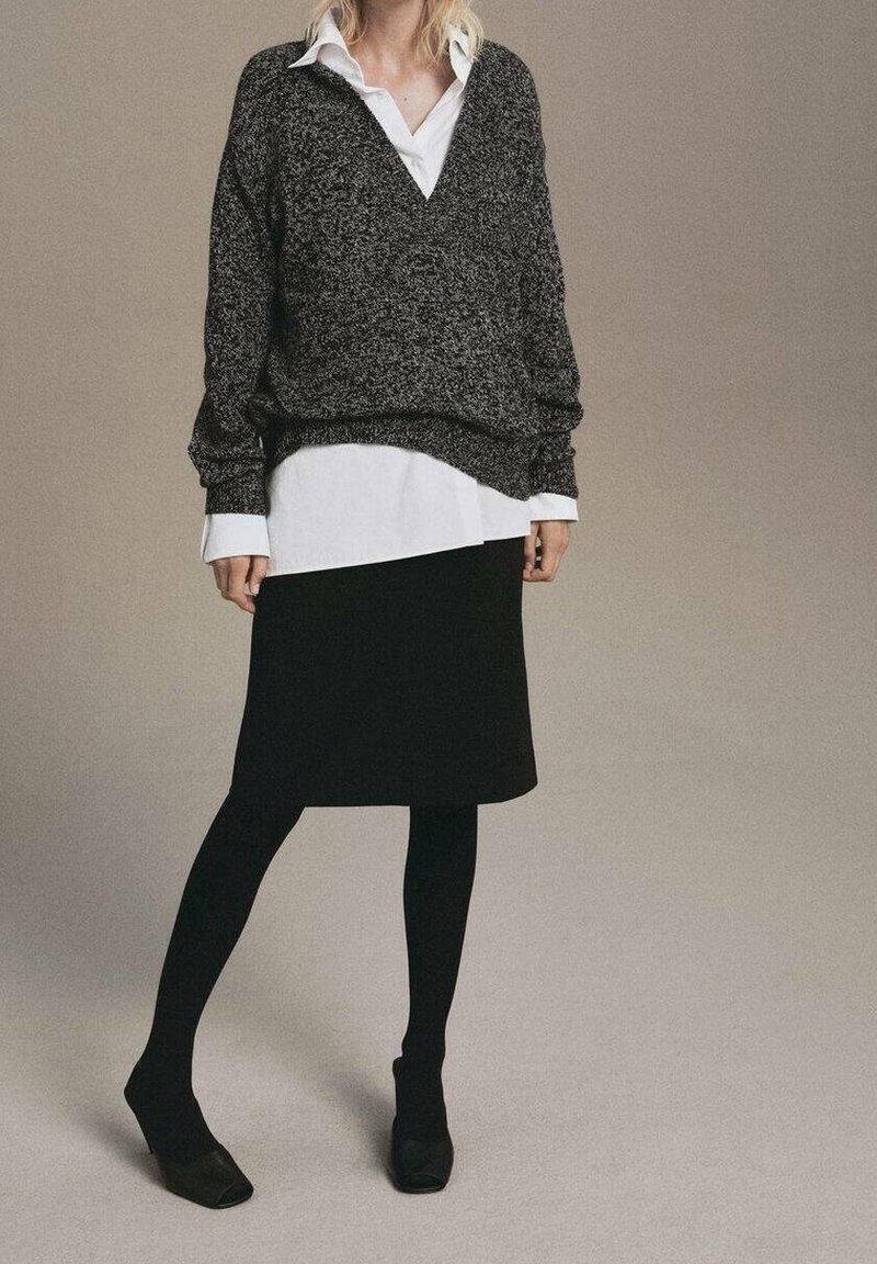 Personne portant une jupe noire, des collants noirs, des chaussures noires, une chemise blanche à col sous un pull gris ample à col en V, debout devant un fond uni.