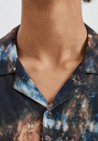 Kurzarmhemd mit einem blauen und braunen abstrakten Muster. Hergestellt aus glattem Stoff, verfügt es über einen eingekerbten Kragen und einen einzelnen Knopfverschluss.