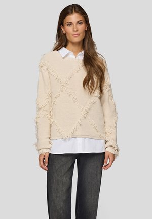 Beige sweater with fringe accents in a diamond pattern. Worn over a white shirt, featuring long sleeves and a relaxed fit.