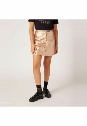 Kobieta ubrana w błyszczącą beżową mini spódniczkę na guziki, czarny T-shirt z napisem, czarne siateczkowe skarpety i czarne masywne mokasyny, stojąca na jednolitym tle.