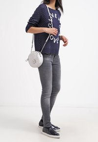 Marinblå tröja med vit text, grå tajta jeans och en rund silverfärgad axelväska med kedjesnodd. Svarta slip-on skor med texturerad detalj.