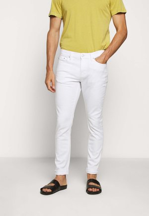 Hombre vestido con jeans blancos ajustados, camiseta amarilla de manga corta y sandalias negras tipo slide, de pie con una mano en el bolsillo.