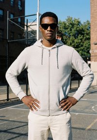 Jeune homme portant des lunettes de soleil et un sweat à capuche gris clair, debout les mains sur les hanches sur un terrain de basket en plein air.