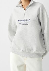 Grauer Fleece-Pullover mit einem Viertel-Reißverschluss am Kragen, einem blauen aufgedruckten "MARSEILLE" Logo und Text auf der Brust, mit gerippten Bündchen und Saum.