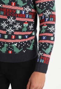 Pull en tricot avec un fond bleu marine orné de dinosaures verts, de flocons de neige et du texte "Joyeux Rex-mas" en rouge. Poignets et ourlet côtelés.