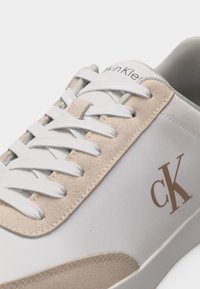 Κοντινό πλάνο σε λευκό και μπεζ αθλητικό παπούτσι με λευκά κορδόνια, σουέτ λεπτομέρειες και καφέ λογότυπο "cK" στο πλάι.