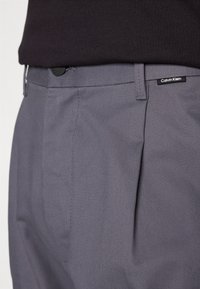 Pantaloni grigi su misura con frontale piatto, caratterizzati da una texture sottile e un'etichetta nera di Calvin Klein sulla vita.