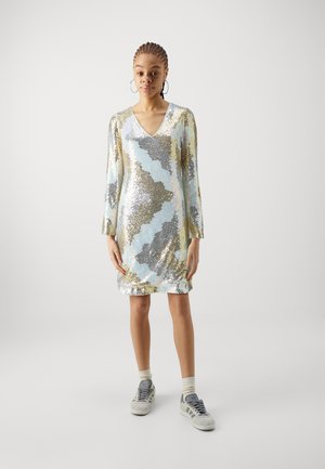 Robe pailletée avec un décolleté en V, des manches longues et un motif en zigzag dans des tons argent, or et bleu. Portée avec des baskets grises et des chaussettes.