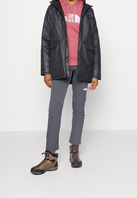 Czarna izolowana kurtka z kapturem, różowy sweter, szare spodnie i brązowe buty trekkingowe. Widoczne logotypy znanych marek na odzieży.