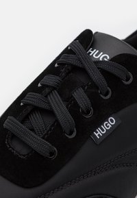 Gros plan d’une basket noire avec des lacets épais noirs, une tige en daim et tissu, et un logo blanc « HUGO » sur la languette et l’étiquette latérale.