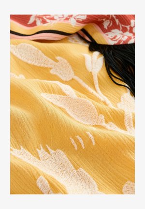 Tissu jaune avec un motif floral abstrait en blanc. Présente un léger éclat et une texture douce, avec des franges noires le long d'un bord.