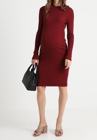 Robe en maille côtelée bordeaux à manches longues, silhouette ajustée, longueur au genou. Portée avec un sac à main noir et des sandales à talons carrés rose pâle.