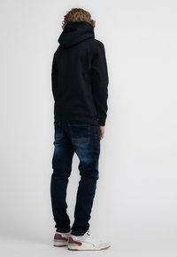 Felpa con cappuccio nera, jeans e scarpe bianche con dettagli bordeaux. Vista posteriore semplice, che mostra il tessuto testurizzato della felpa.