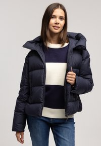Granatowa kurtka puchowa z odpinanym kapturem, posiadająca pikowane elementy, zamek błyskawiczny z przodu oraz boczne kieszenie; noszona na paskowanym swetrze.