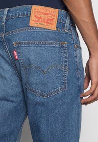 Vaqueros de denim azul con una etiqueta de logo roja visible en el bolsillo trasero, costuras doradas y un parche de cuero marrón claro con detalles de la marca.