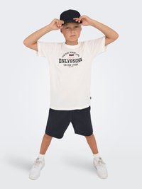 White t-shirt with black print, black shorts, white sneakers, and a black cap. The outfit features a relaxed fit and casual style.