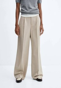 Personne portant un pantalon beige à jambes larges, un haut gris à manches courtes et des mocassins bicolores, debout devant un fond blanc uni.