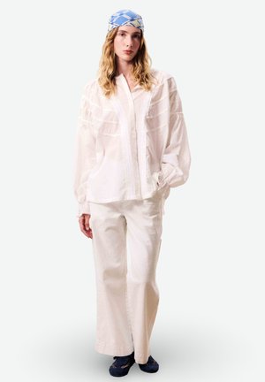 Jeune femme portant un chemisier blanc ample, un pantalon blanc, un foulard bleu à motifs et des chaussures décontractées bleu foncé, debout devant un fond uni.