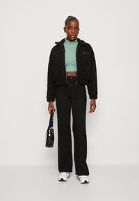 Sort cropped fleecejakke med lynlås, grøn strikket sweater, højtaljede sorte flairbukser og hvid-sorte sneakers. Holder en sort taske.