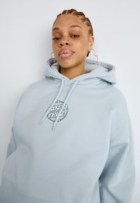 Sweatshirt azul claro com um bolso canguru, capuz com cordão e um logotipo bordado em forma circular que apresenta texto e estrelas no peito.