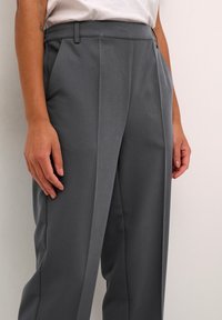 Pantalons taillés gris avec une texture lisse, taille haute, plis sur le devant et poches latérales, associés à une chemise blanche.