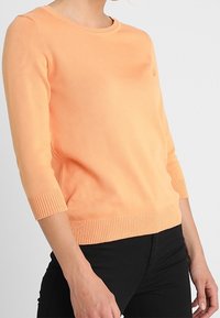 Pull-over léger orange avec un col rond, des poignets et un ourlet côtelés. Fabriqué dans un matériau doux avec une texture lisse.