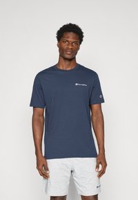 Dunkelblaues T-Shirt aus Baumwolle mit kleinem weißen Logo auf der linken Brust und einem Logodetail am Ärmel, kombiniert mit grauen Shorts.
