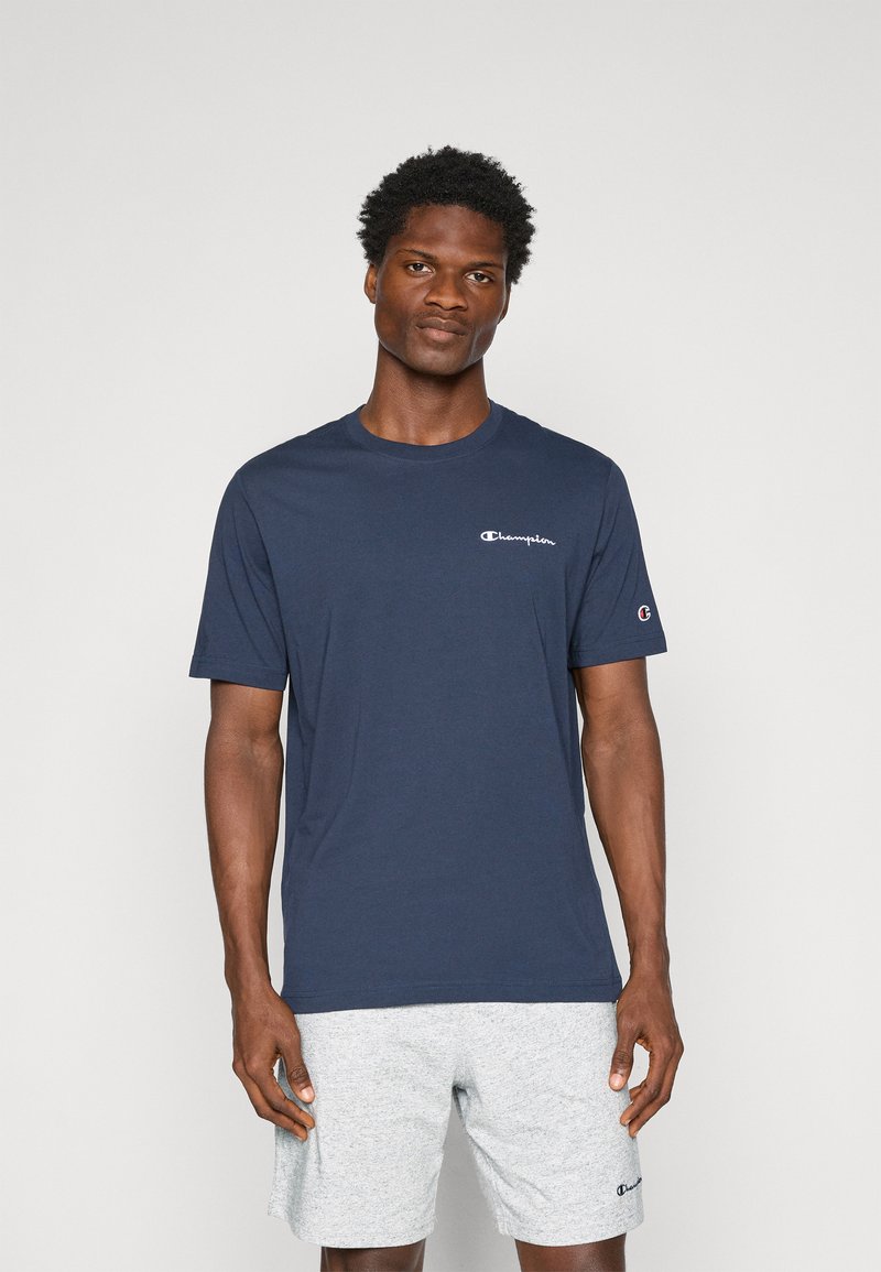 Dunkelblaues T-Shirt aus Baumwolle mit kleinem weißen Logo auf der linken Brust und einem Logodetail am Ärmel, kombiniert mit grauen Shorts.