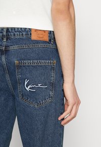 Jeans i mörkblå denim med en strukturerad yta. Har en läderlapp vid midjan och en broderad logotyp på bakfickan.