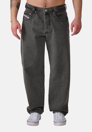 Mann trägt locker sitzende schwarze Jeans und weiße Turnschuhe, steht mit entspannten Armen an den Seiten vor einem einfarbigen Hintergrund.