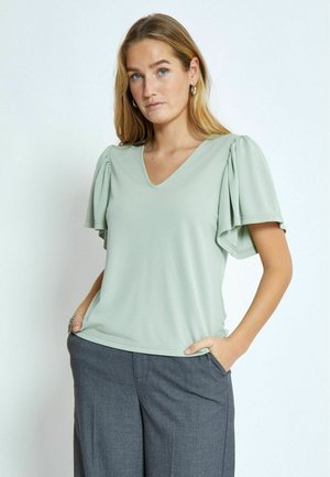 Light green v-neck blouse with flutter sleeves, featuring soft fabric and subtle texture; paired with gray trousers, hands in pockets.