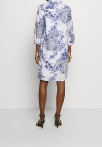 Donna che indossa un vestito bianco fino al ginocchio con intricate ornamentazioni blu, maniche a lunghezza media e sandali neri con tacco alto, in piedi di spalle.