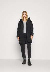 Schwarze Daunenjacke mit Kapuze, mit einem gesteppten Design, vorderen Knöpfen und Taschen. Kombiniert mit einem grauen Pullover und schwarzen Jeans.