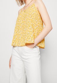 Camiseta sin mangas amarilla con flores, que presenta pequeñas flores blancas y moradas en un corte holgado, combinada con pantalones anchos blancos.