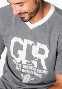 Graues T-Shirt mit kurzen Ärmeln und weißem V-Ausschnitt, versehen mit markanter, abgenutzter "GDR" Grafik und dem Text "Athletic Sporting Goods", der auf die Vorderseite gedruckt ist.