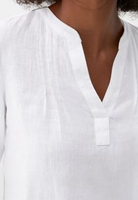 Weiße Leinenbluse mit V-Ausschnitt und dezenten Steppdetails entlang des Ausschnitts, die eine leichte und atmungsaktive Textur zeigt.