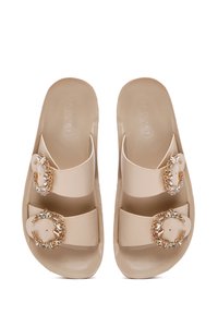 Nude PVC slide sandals featuring two wide straps adorned with circular gold hardware and clear gems, with a smooth, matte finish.
