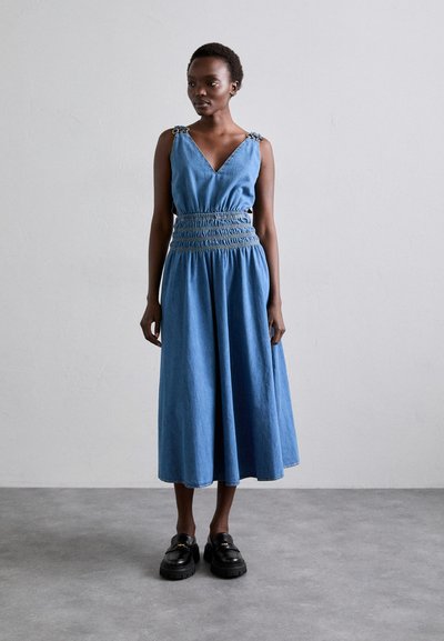 Rochie din denim cu decolteu în V, talie striată și bretele accentuate cu lanț; lungime până la genunchi, cu o croială lejeră și textură moale. Pantofi loafers masivi negri purtați.