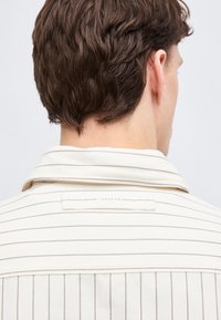 Άνδρας με κοντά καστανά μαλλιά που φοράει κρεμ πουκάμισο με λεπτές σκοτεινές οριζόντιες και κάθετες ρίγες, εμφανίζεται από πίσω και ελαφρώς στραμμένος προς τα δεξιά.