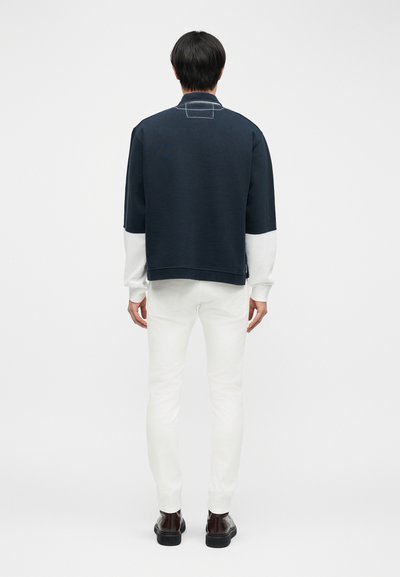 Άτομο που στέκεται με την πλάτη, φοράει μπλε ναυτικό και λευκό μπλουζάκι μακρυμάνικο, λευκό παντελόνι και σκούρα καφέ παπούτσια σε λευκό φόντο.