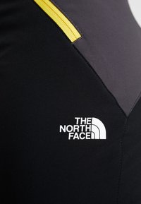 Musta tekstiilivaatte, jossa on keltainen vetoketju ja valkoinen "The North Face" -logo, joka on näkyvästi esillä pinnalla.