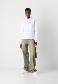Hanorac alb cu glugă, blugi oversized albaștri deschiși și adidași albi, ținând o geantă metalică. Textură netedă, croială lejeră.