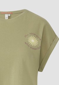Olijfgroene katoenen T-shirt met korte opgerolde mouwen; voorzien van een lichtgele madeliefje grafiek en de tekst "VUL JE DAGEN MET ZONNESCHIJN."
