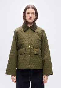 Νεαρή γυναίκα που φοράει πράσινο μπουφάν με καπιτονέ ύφασμα, μεγάλο γιακά και χρυσά κουμπιά, στέκεται μπροστά σε ένα απλό ανοιχτό φόντο.