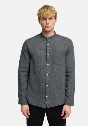 Ung mand med blondt hår iført en tekstureret grå skjorte med mandarinkrave, stående mod en ensfarvet lys baggrund.