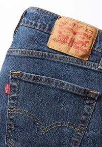 Levi's siniset denim-farkut, joissa on nahkapaneeli punaisella painatuksella, näkyvää ompelua ja punainen etiketti takataskussa. Klassinen viiden taskun muotoilu.