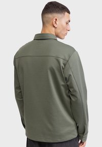 Camisa de manga larga verde oliva con cuello estructurado, corte recto y puños abotonados. Textura suave con detalles de costura mínimos.