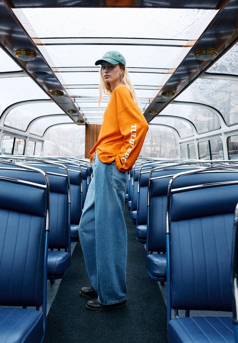 Jeune personne portant un sweat-shirt orange et une casquette verte se tient dans l'allée d'un bus vide au toit en verre avec des sièges bleus.