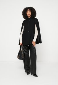 Μαύρη κάπα από μάλλινο μείγμα με ψηλό γιακά και κούμπωμα με ένα κουμπί, φορεμένη πάνω από ένα κρεμ πουλόβερ· συνδυασμένη με μαύρο δερμάτινο παντελόνι και τσάντα tote.